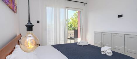 A hotel room with a bed, a hanging light fixture, a view through the window, and a radiator.