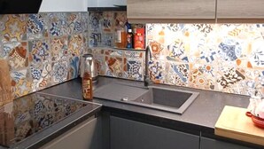 Une cuisine avec un plan de travail noir, un évier et un crédence ornée de carreaux colorés. On y trouve des placards au-dessus du plan de travail et une étagère en bois sur laquelle sont posés divers objets.