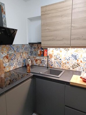 A kitchen with a black countertop, a sink, and a backsplash with colorful tiles. There are cabinets above the countertop and a wooden shelf with items on it. - Apartment Snjezana (Senj)