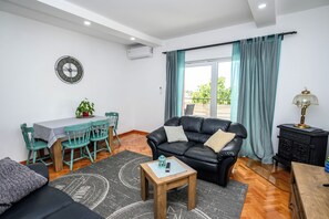 Ein Wohnzimmer mit einer Couch, einem Couchtisch, einem Esstisch mit Stühlen, einer Uhr und Blick durch ein Fenster mit Vorhängen.
