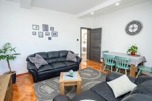 A modern living room with a black leather sofa, a small wooden table, a dining area with a table and chairs, and a wall clock. - Apartment Snjezana (Senj)