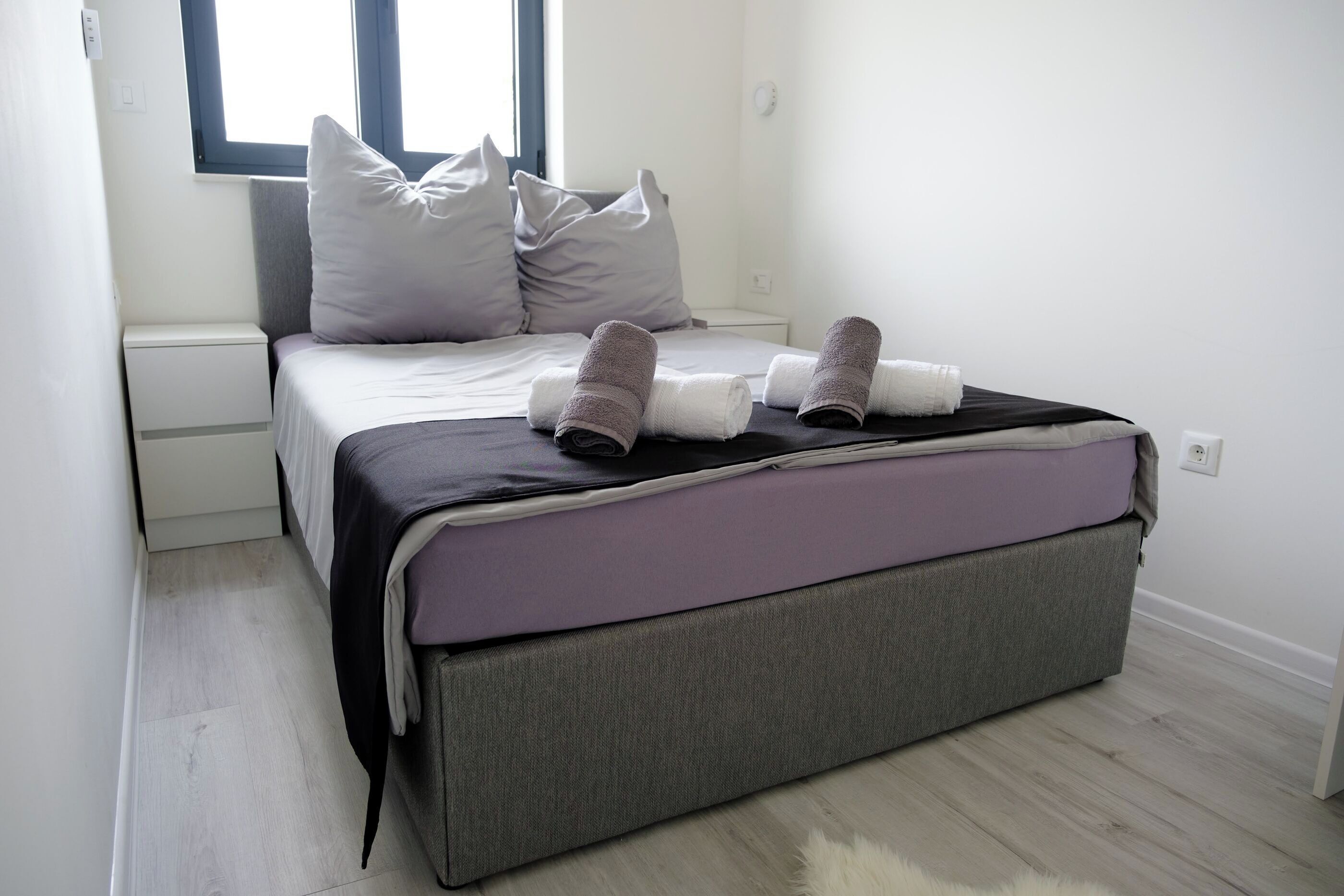 A neatly made bed with pillows and rolled towels, a bedside table, and a window with blinds.