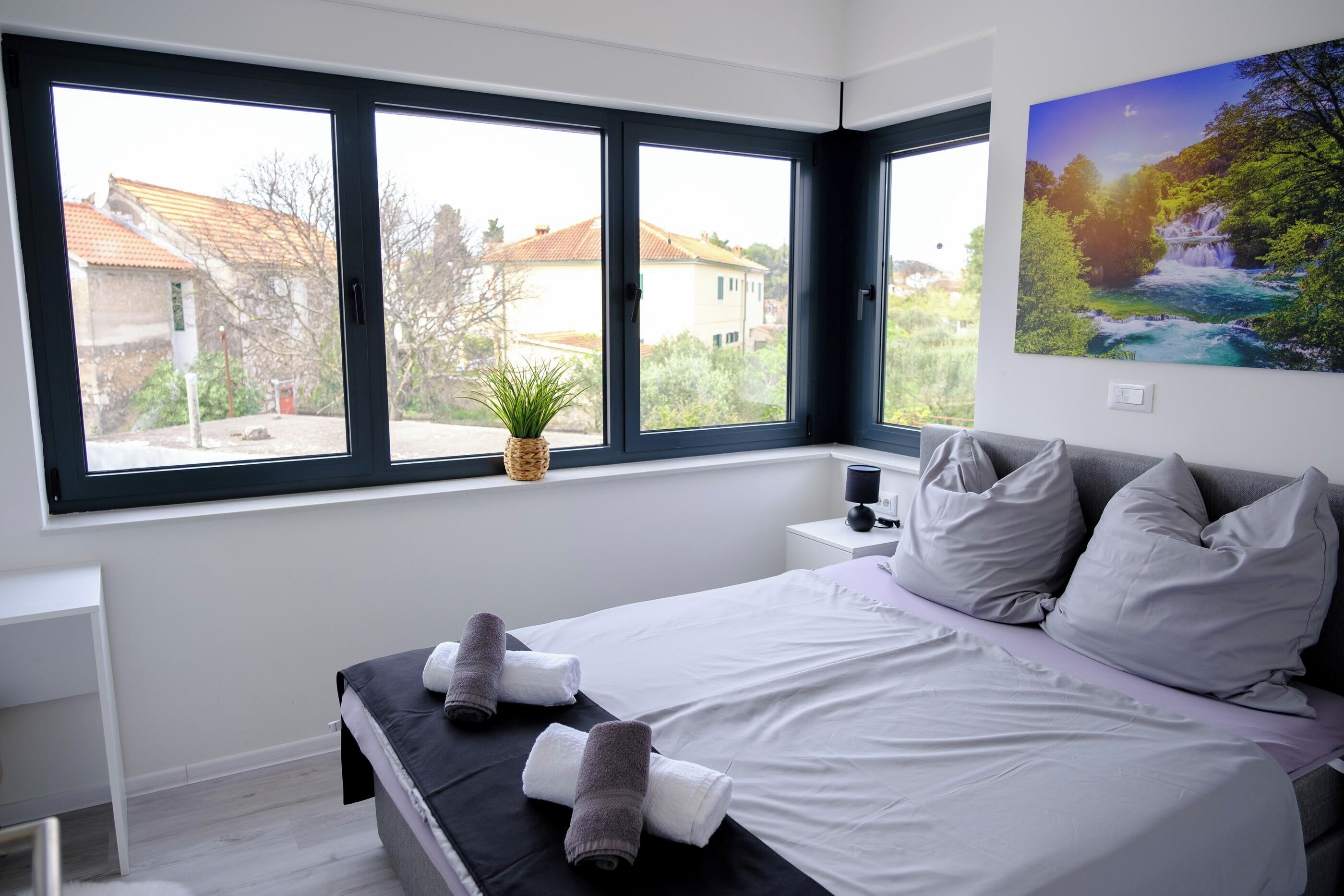 Un dormitorio moderno con una cama grande, dos mesitas de noche, una ventana con vistas a casas y un cuadro en la pared.