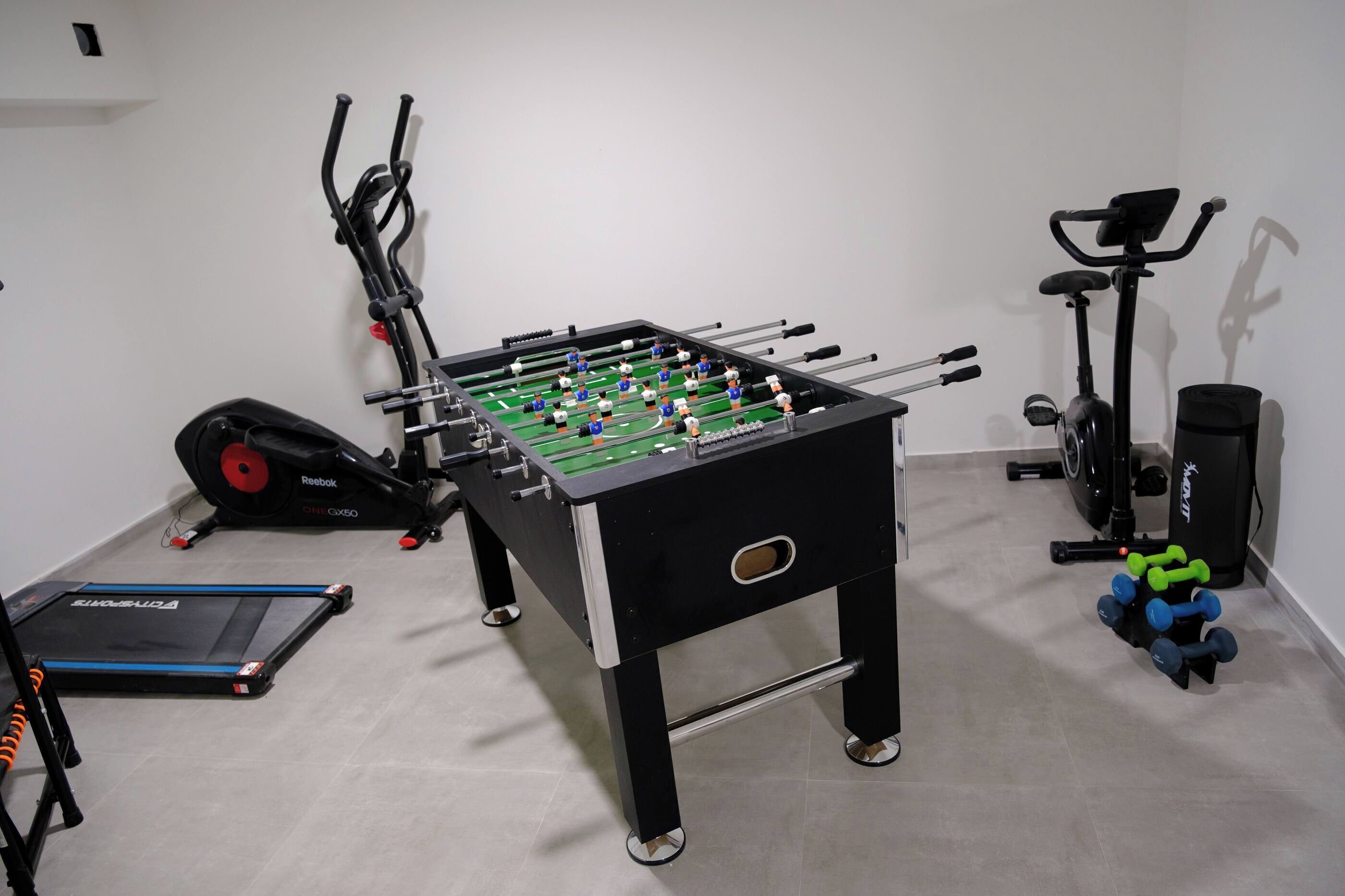 Ein Zimmer mit Kicker, Ellipsentrainer von Reebok, Fahrradergometer und Kurzhanteln.