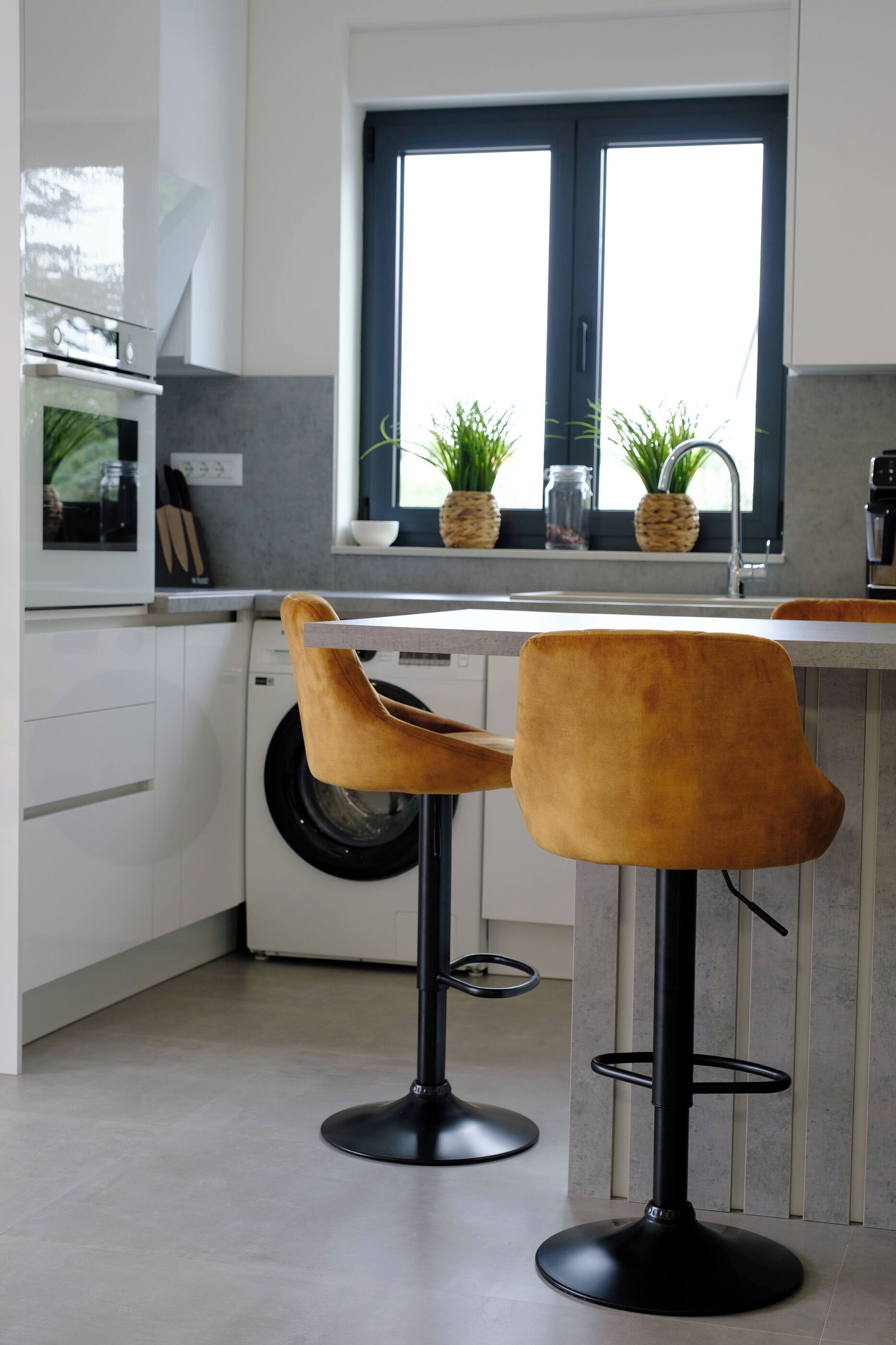 A modern kitchen with a washing machine, two bar stools, and a window with potted plants.