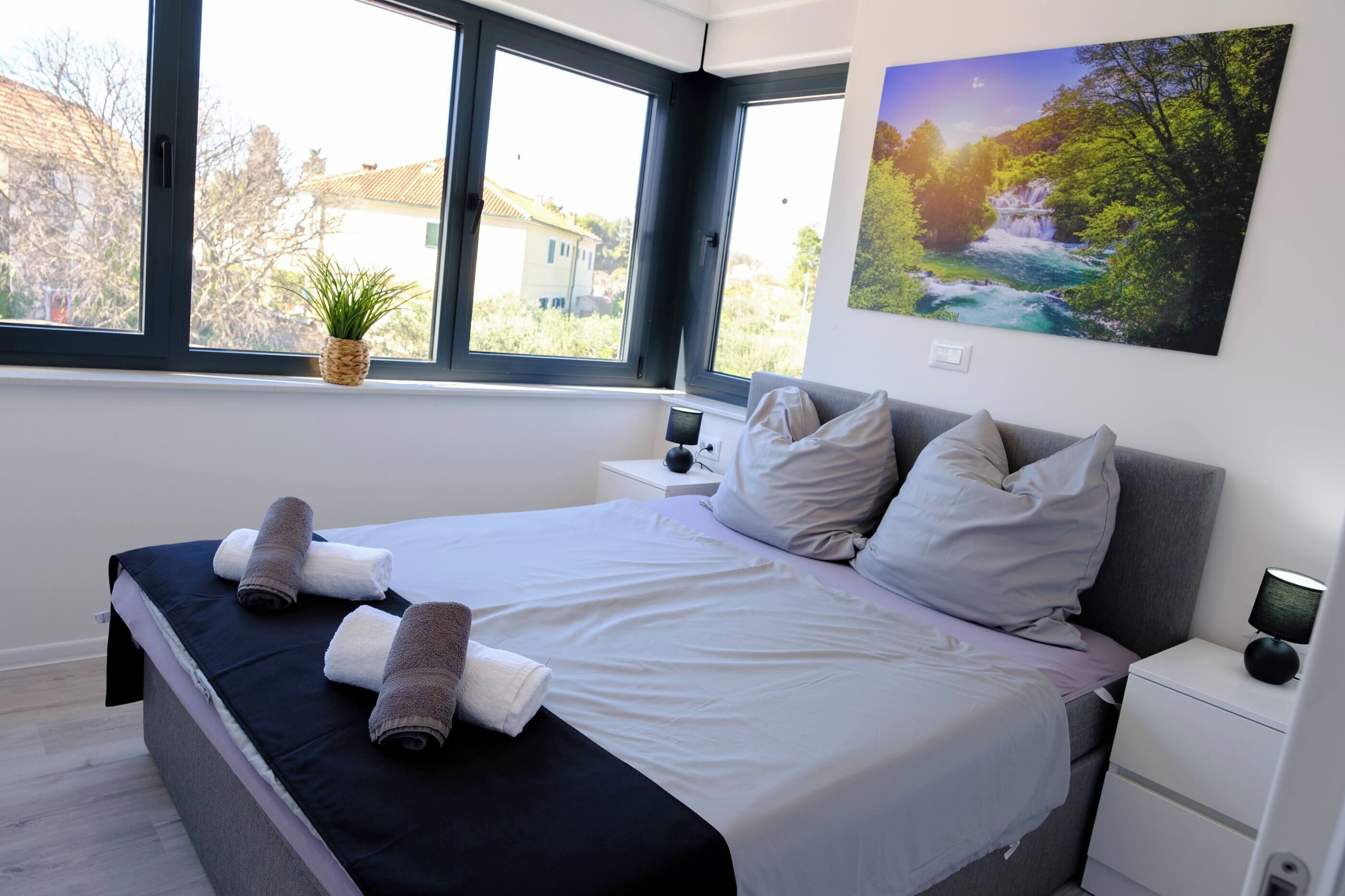 A modern bedroom with a large bed, two bedside tables, a wall-mounted TV, and a window with a view of a house and trees.