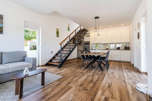 Een moderne keuken met witte kasten, een eethoek met een houten tafel en stoelen, een trap met houten leuning, en een grijze bank in een woonruimte.