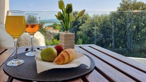 A table with two glasses of a yellow beverage, a croissant, and two apples on a placemat, with a vase of tulips and a view of greenery and buildings. - Villa Raslina Sea View (Sibenik)