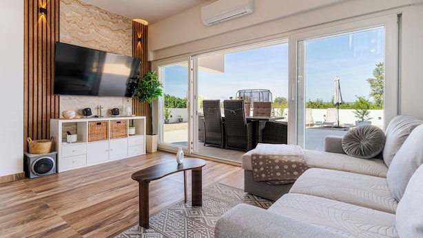 Ein modernes Wohnzimmer mit Flachbildfernseher, Wandverkleidung aus Holz, Schiebeglasfenstern zu einer Außenpatio und einem Sofa mit gemusterten Kissen.