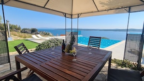 A wooden table with two chairs, a glass of wine, and a fruit bowl under a canopy overlooking a swimming pool and a scenic view of the sea and hills. - Villa Dream Come True (Jelsa)