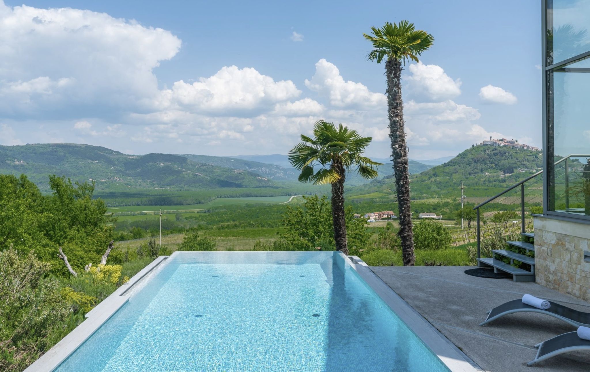 A swimming pool with clear water, surrounded by lush greenery and palm trees, overlooking a scenic landscape with hills and a distant village.
