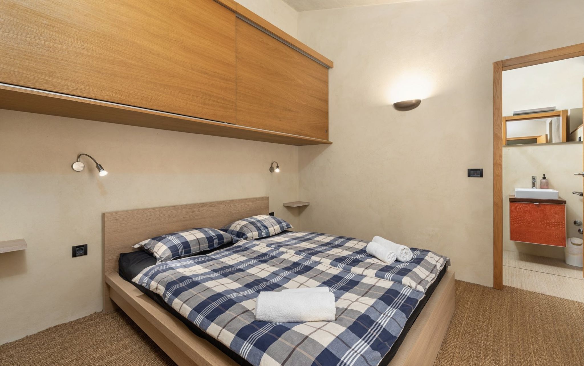 Een slaapkamer met een bed, een houten hoofdbord, een nachtkastje, een wandlamp en een zichtbare badkamer door een open deur.