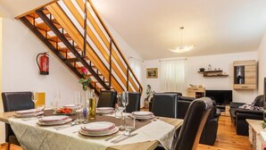 Uma área de refeições com escada de madeira, uma mesa de jantar posta para quatro pessoas e uma sala de estar com sofá e televisão.