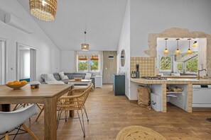 Uma cozinha moderna com ilha central, área de jantar com cadeiras de vime e sala de estar com sofá.