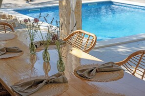 Uma área de refeições à beira da piscina com mesa de madeira, jogos americanos trançados e cadeiras de vime.