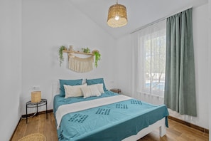 Ein Schlafzimmer mit einem Bett, einer Pendelleuchte, einem Fenster mit Vorhängen und einem an der Wand befestigten Regal mit Pflanzen.