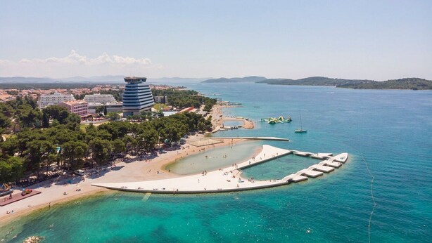 Vista aerea di una zona costiera con spiaggia, una torre e un porto turistico.