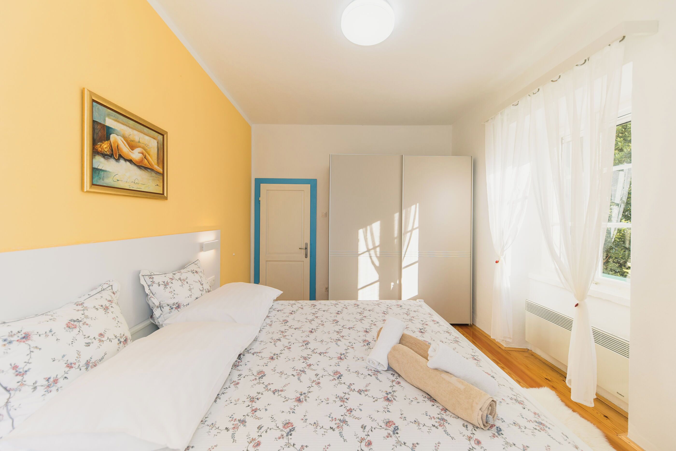 A bedroom with a bed, a painting on the wall, a wardrobe, and a window with curtains.