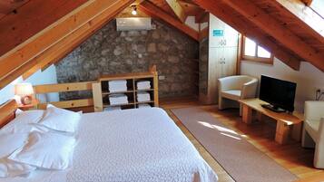 A cozy attic bedroom with a bed, a television, a stone wall, and wooden beams.
