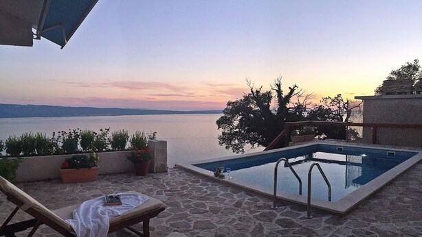 A poolside area with lounge chairs, a small table with a book, and a view of the sea at sunset. - Villa Peace (Dugi Rat)