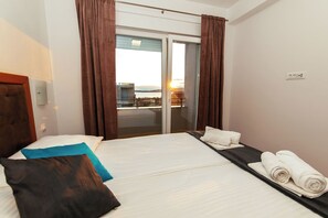 Una camera d'hotel con un letto, un piccolo tavolino con asciugamani e una porta scorrevole in vetro che dà su un balcone, con vista sul tramonto.