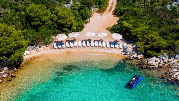 Une vue aérienne d’une plage aux eaux turquoise et limpides, bordée d’une rangée de transats et où un bateau est amarré près du rivage.