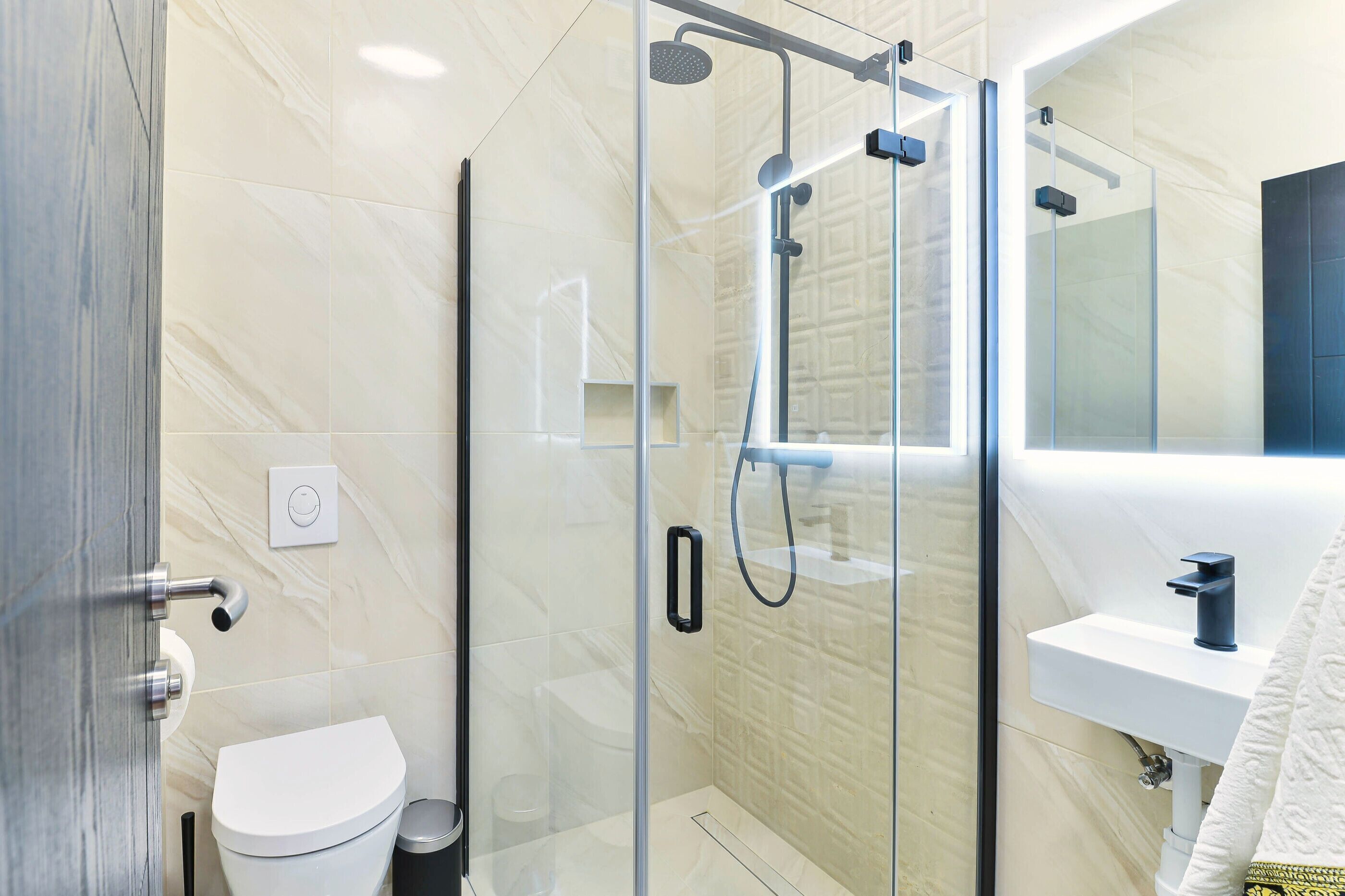 Une salle de bain moderne avec une douche à parois de verre, des toilettes blanches et un lavabo surmonté d’un miroir.