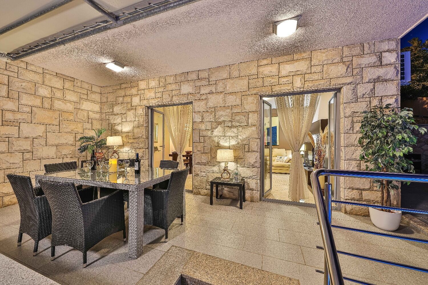 A dining area with a stone wall, a glass table, wicker chairs, and a small side table with a lamp.