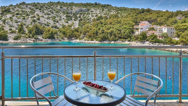 Ein Balkon mit Tisch, zwei Stühlen und Blick auf das türkisfarbene Meer sowie eine Hügelkette mit Gebäuden.