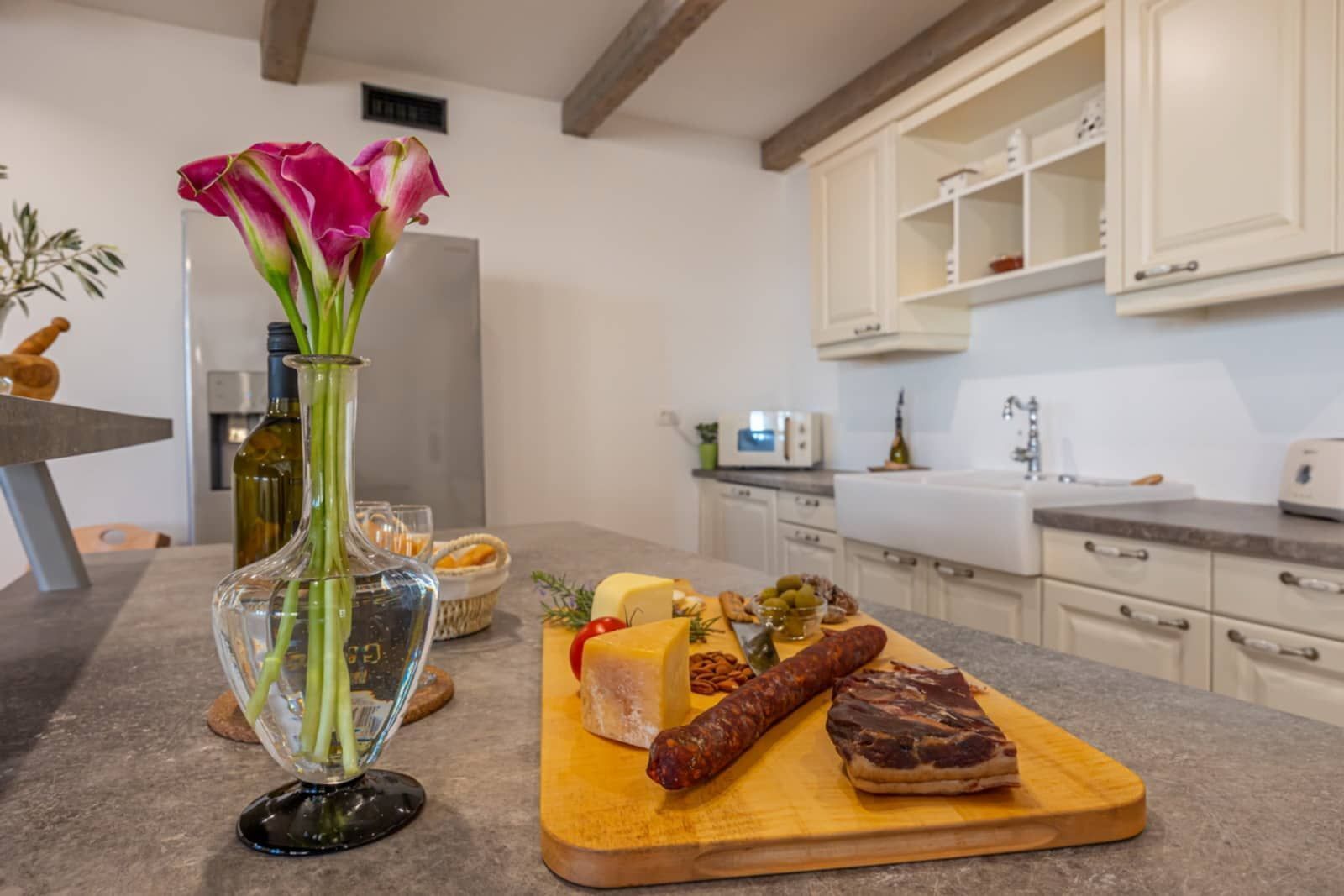 A kitchen with a marble countertop, a vase with flowers, and a variety of food items including cheese, cured meats, and pickles.