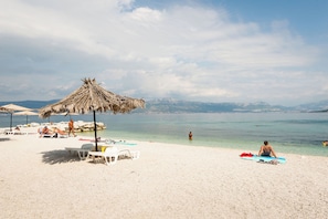 Ein Strand mit weißem Sand, einem Reetdachschirm und freiem Blick auf das Meer und die dahinterliegenden Berge.