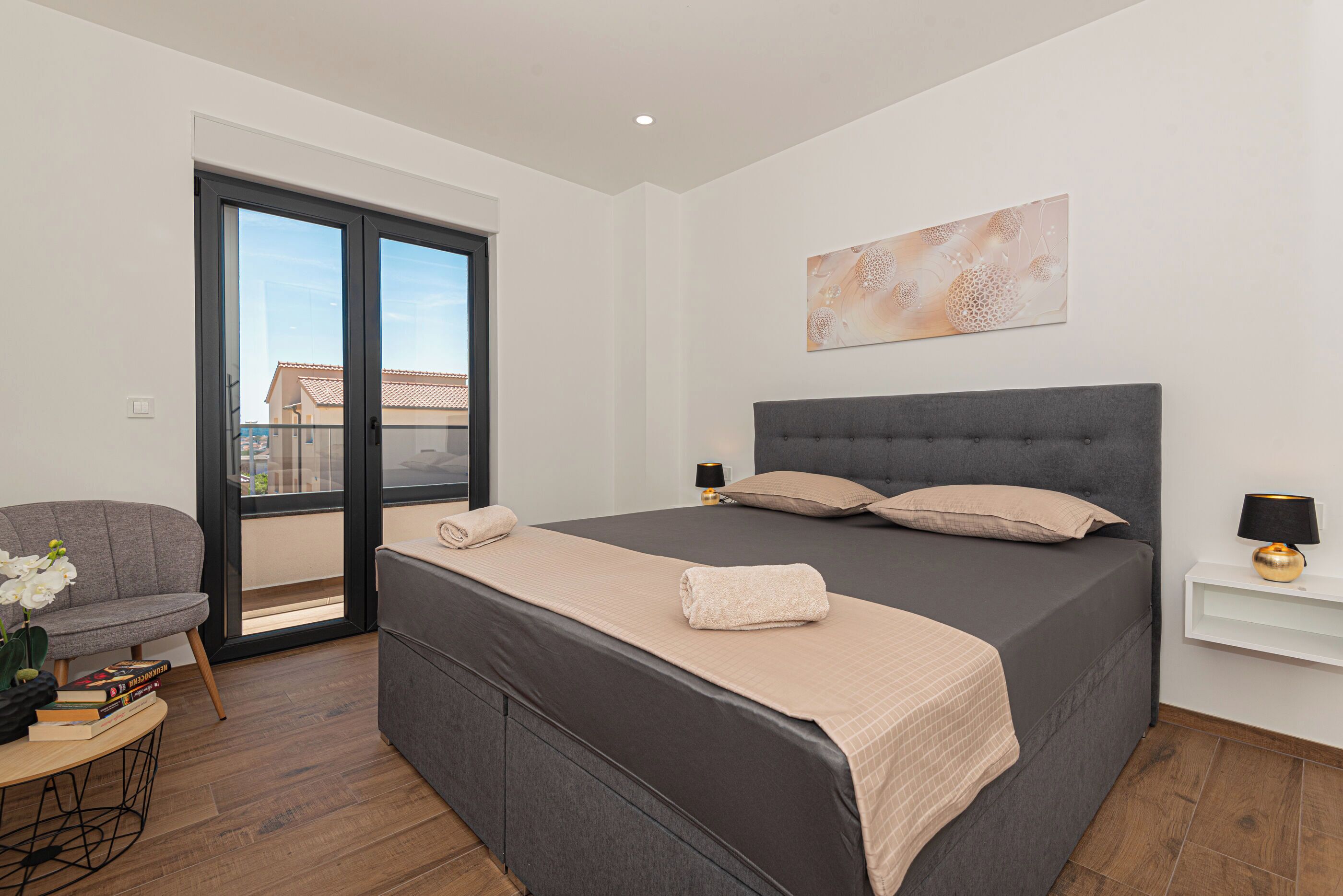 A modern bedroom with a large bed, a grey sofa, a small table with books, and a balcony with a view of buildings.