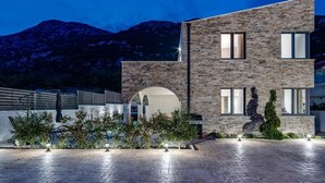 A modern two-story building with a stone facade, an arched entrance, and a paved courtyard with outdoor lighting. - Villa Eden With Heated Pool (Omis)