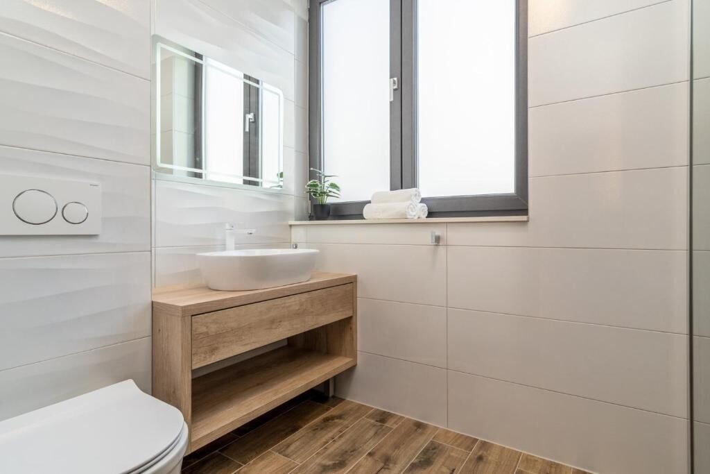 A modern bathroom with a white toilet, a wooden vanity with a white sink, and a window above the sink.