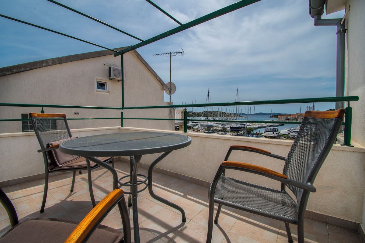 A balcony with a round table and two chairs, overlooking a marina with boats and buildings in the distance.