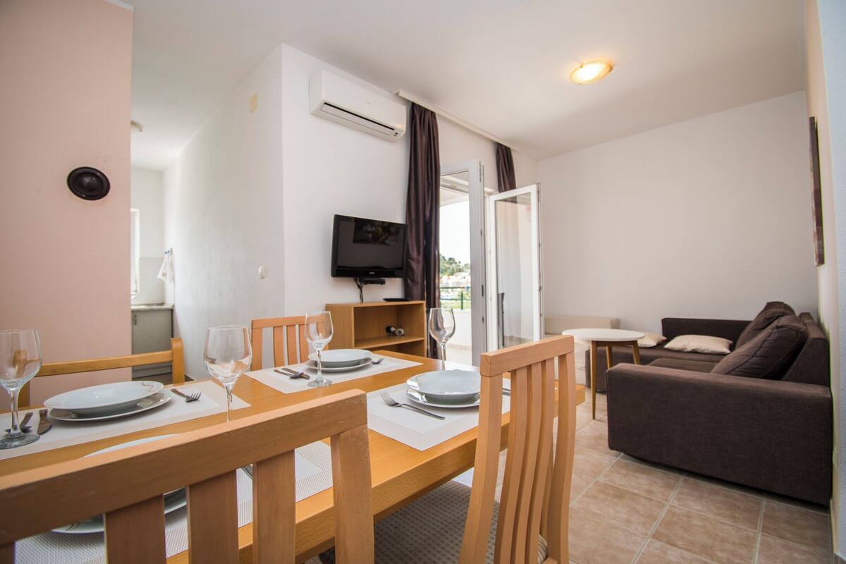 A dining area with a table set for two, a television, and a sofa in the background.