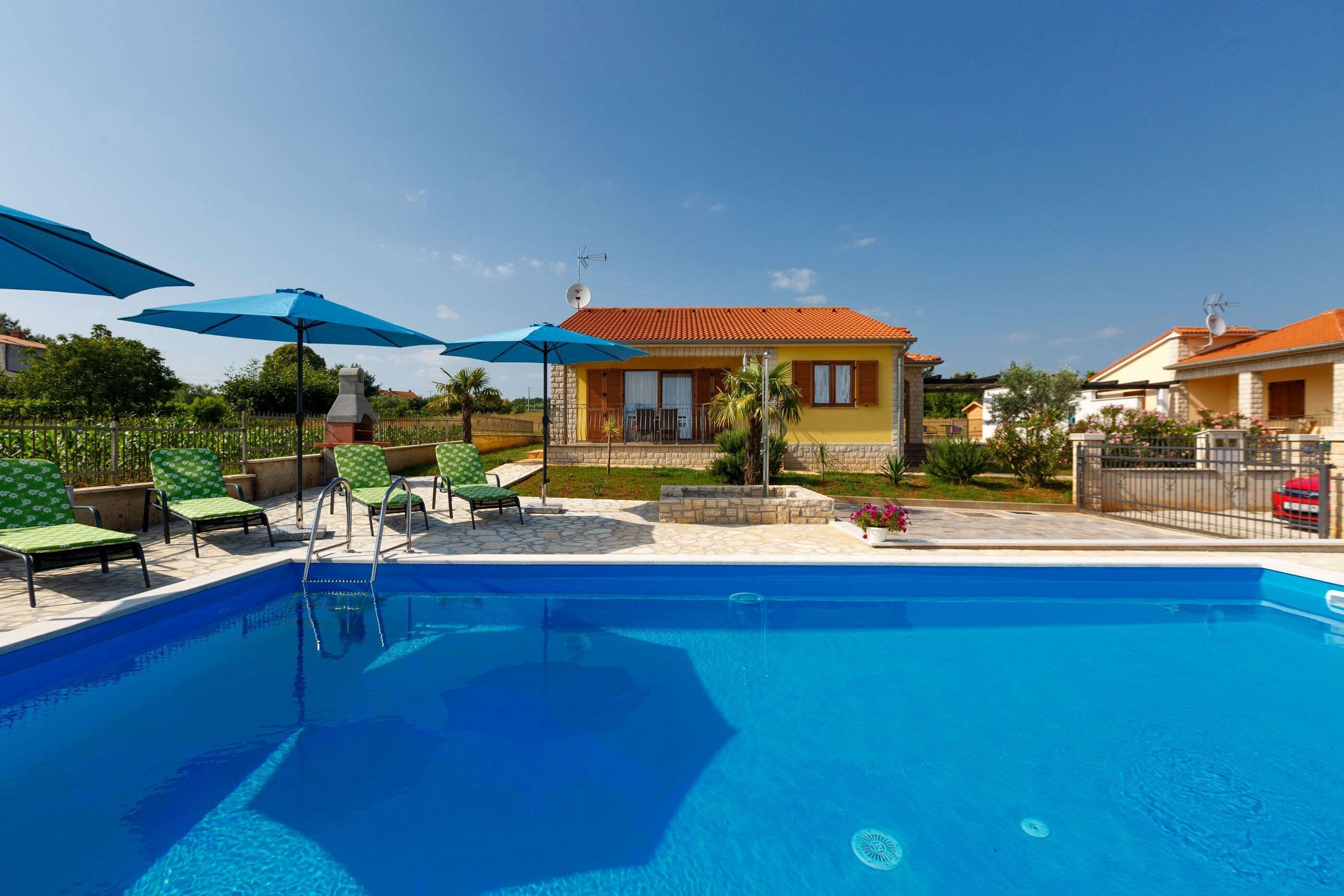 A swimming pool with lounge chairs and umbrellas, surrounded by a house with a red roof and a garden.