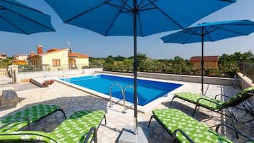 Piscina com espreguiçadeiras verdes e guarda-sóis azuis, em volta de ladrilhos de pedra e um céu claro.