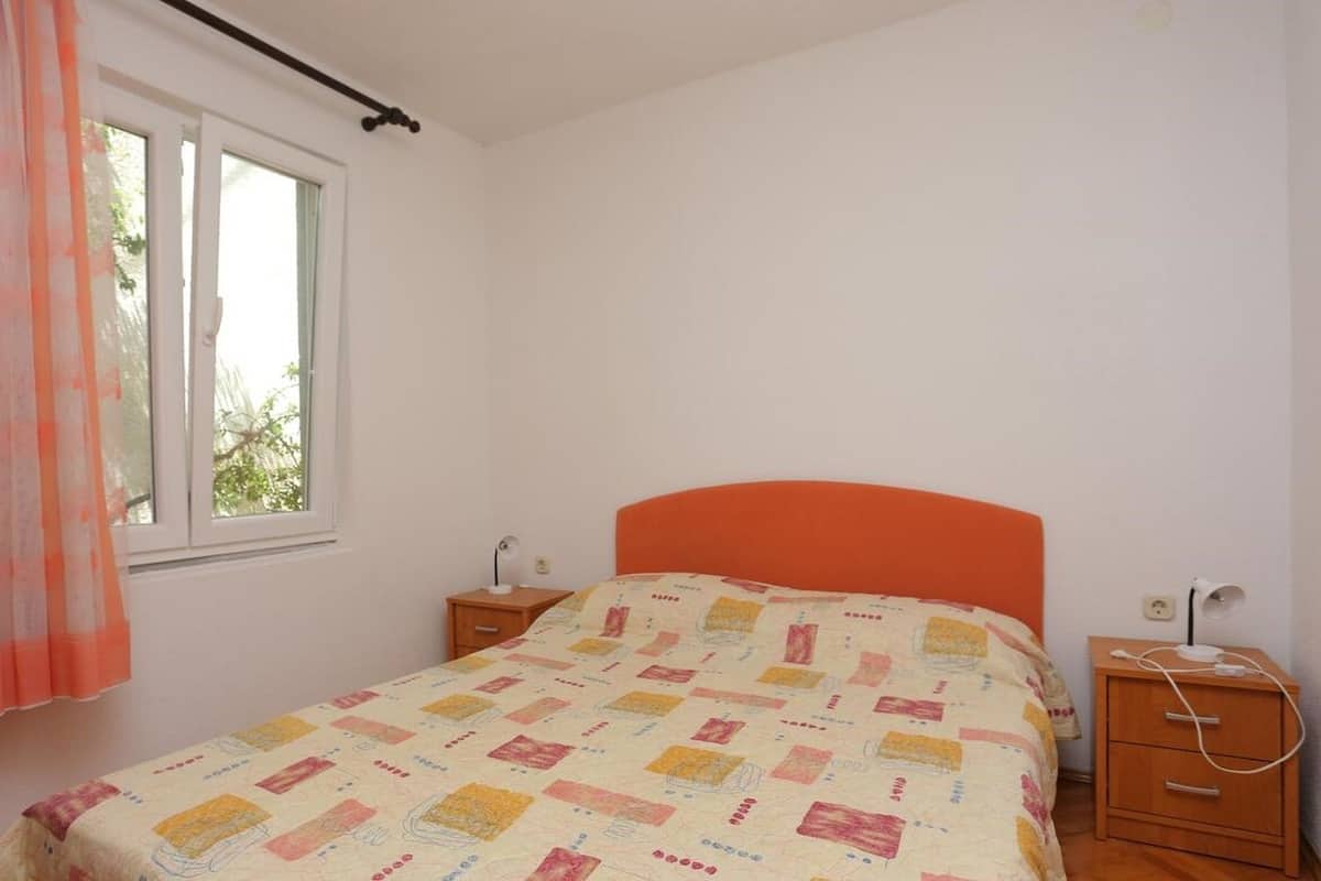 A bedroom with a single bed, a window with curtains, a bedside table with a lamp, and a nightstand with a lamp.