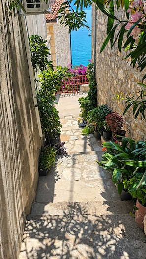 Un estrecho sendero de piedra flanqueado por macetas con plantas que conduce a una puerta roja y vistas al mar.
