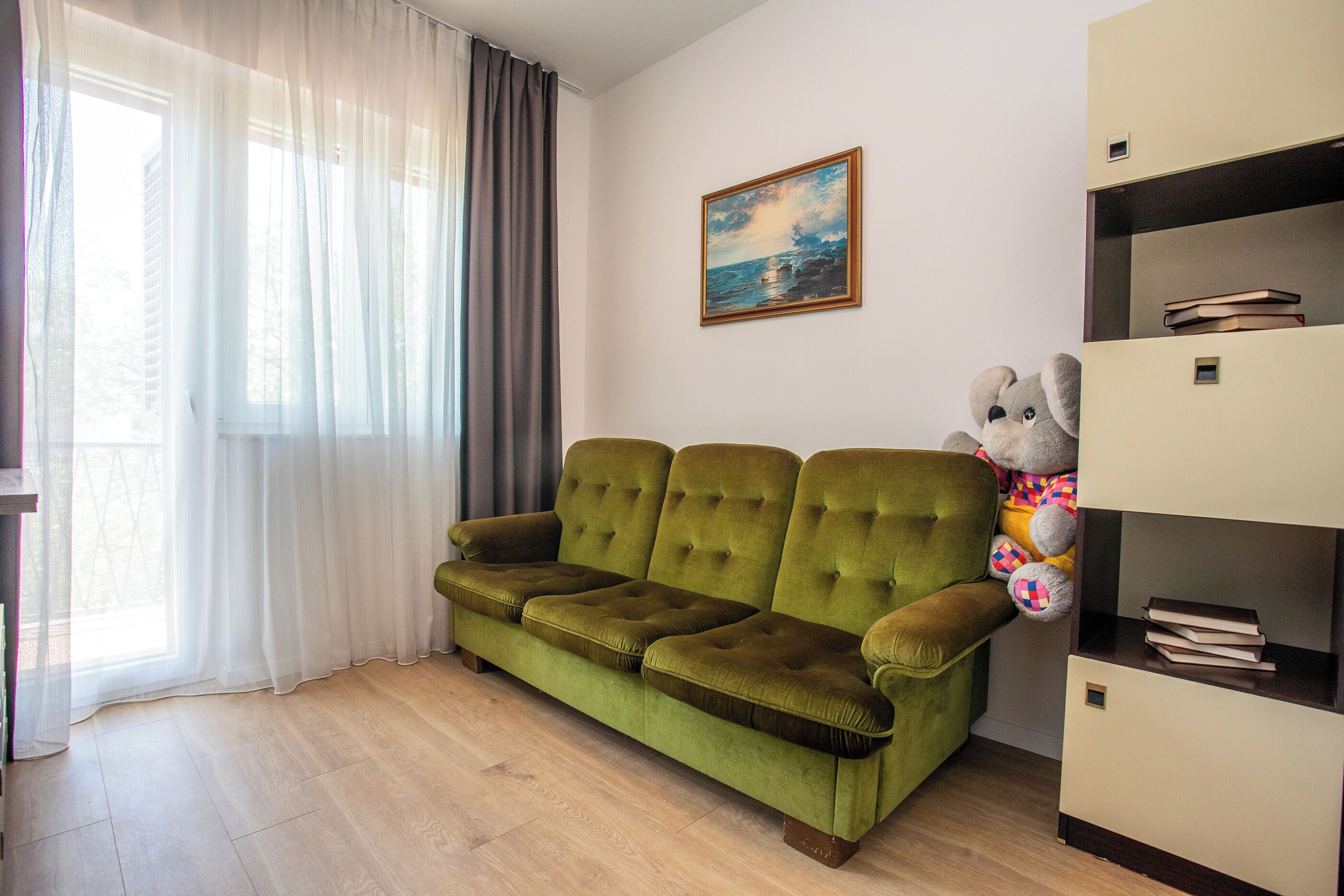 A green sofa, a large stuffed animal, and a bookshelf in a room with a window and a painting on the wall.