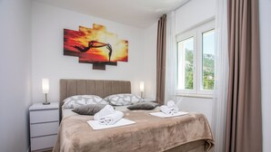 Cama bem arrumada com cobertura bege, duas mesas de cabeceira com luminárias, uma obra de arte na parede e uma janela com cortinas.