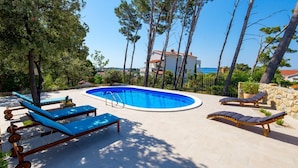 A swimming pool with lounge chairs, surrounded by trees and a stone wall, with a view of the sea and buildings in the background. - Villa Arba (Rab)