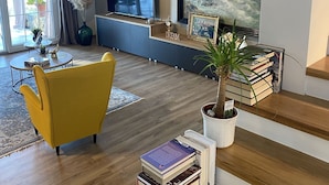 A modern living room with a yellow armchair, a flat-screen TV, a large painting, and a staircase with books. - Villa Arba (Rab)