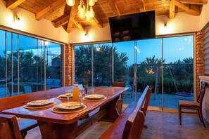 An outdoor dining area with a wooden table set for four, large windows, and a TV mounted on the wall. - Villa Lunis (Vodice)