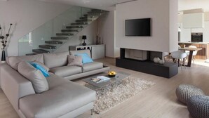 A modern living room with a grey sofa, a coffee table, a fireplace, a flat-screen TV, and a staircase leading to an upper level. - Villa Royal Green (Rasa)
