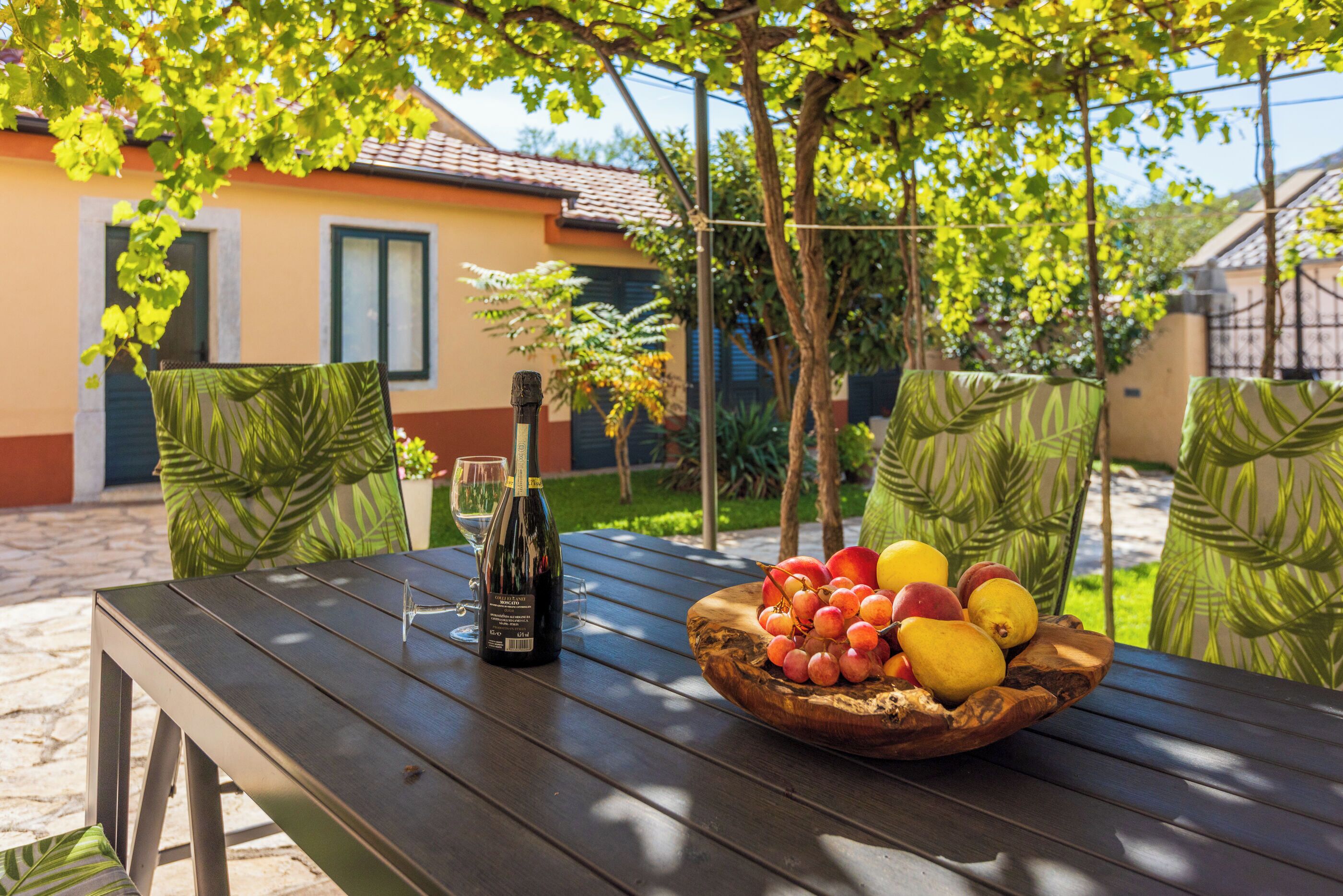 Une table en bois, sur laquelle se trouvent un saladier de fruits et une bouteille de vin, est placée à l’extérieur, agrémentée de coussins verts à motifs, et une maison se profile à l’arrière-plan.