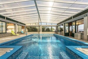 Ein Hallenbad mit Glasdach, klarem blauen Wasser und Liegestühlen.
