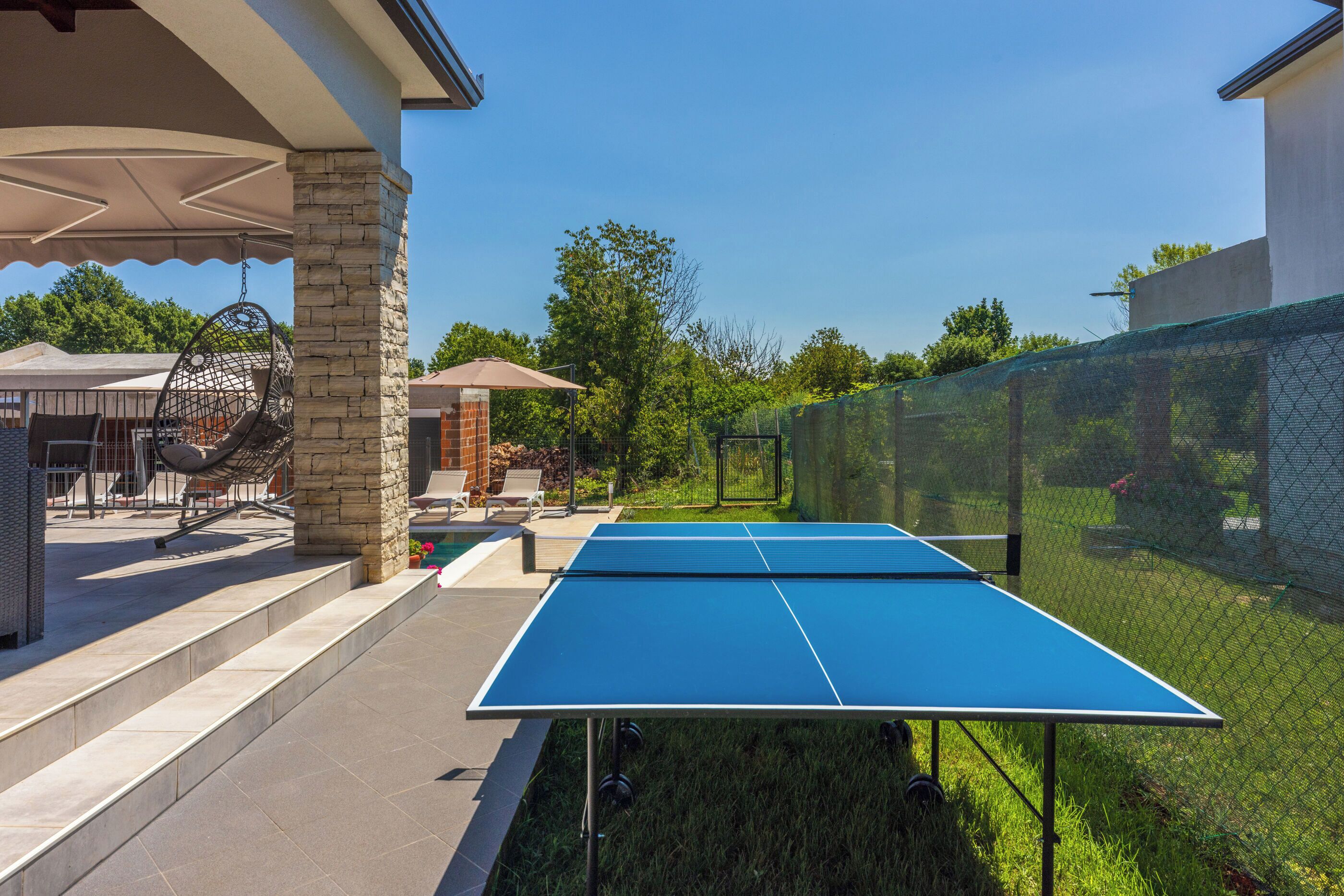 An outdoor ping pong table with a netted enclosure, stone pillars, and outdoor furniture.