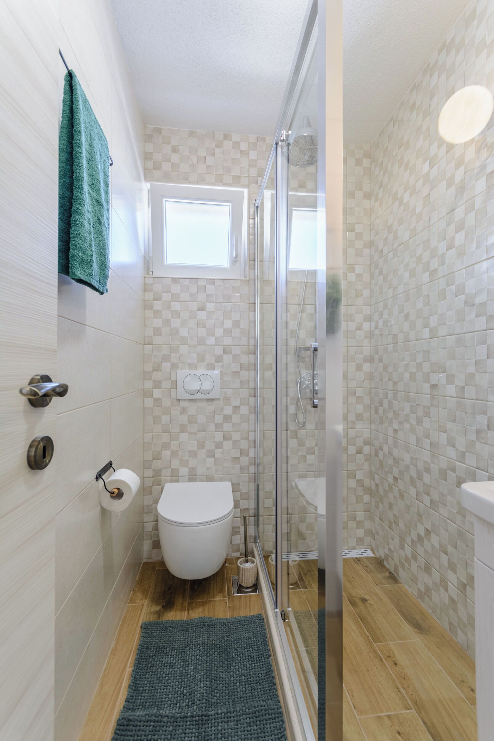 A bathroom with a toilet, a shower with a glass door, a small window, and a green rug.